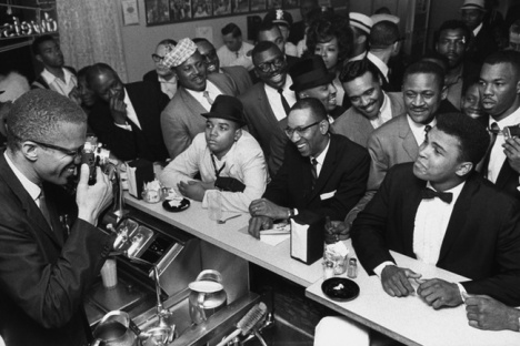 Malcolm X behind the counter at a restaurant, photographing Cassius Clay.